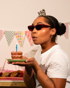 Person holding a stack of cookies with a lit candle, wearing sunglasses and a birthday crown, with a festive background.