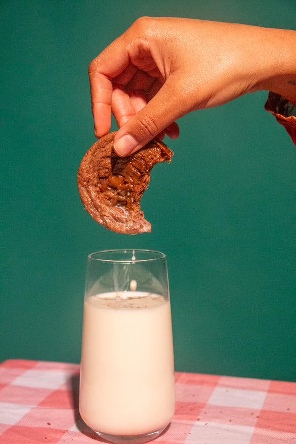 Hand dipping a cookie into a glass of milk on a checkered tablecloth with a green background